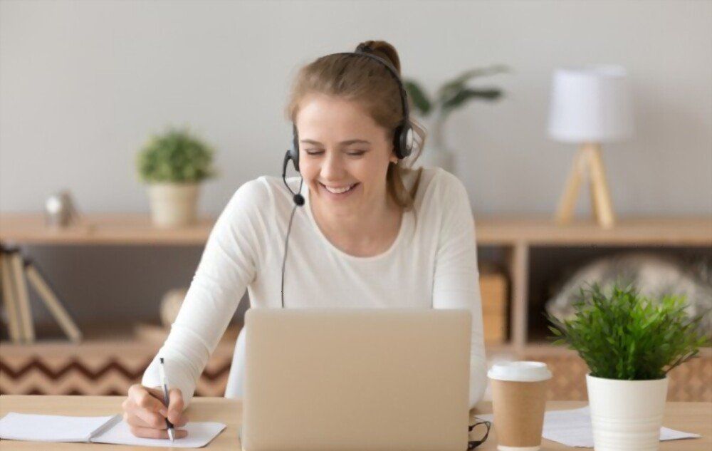 Young woman in a white shirt working on a laptop with a headset on, taking notes.